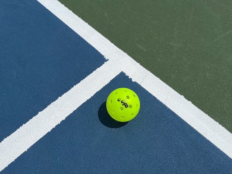 pickleball-on-court A bright yellow pickleball resting on the lines of a blue and green court