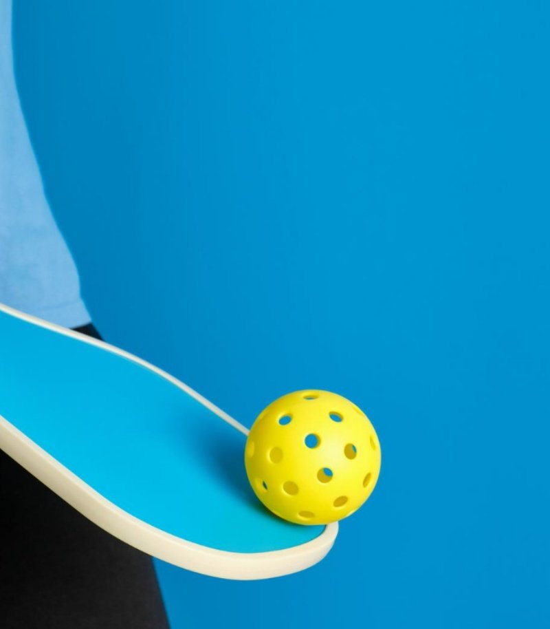 Minimalist pickleball paddle and ball design in vibrant colors Close-up of a blue pickleball paddle balancing a yellow pickleball against a blue background.