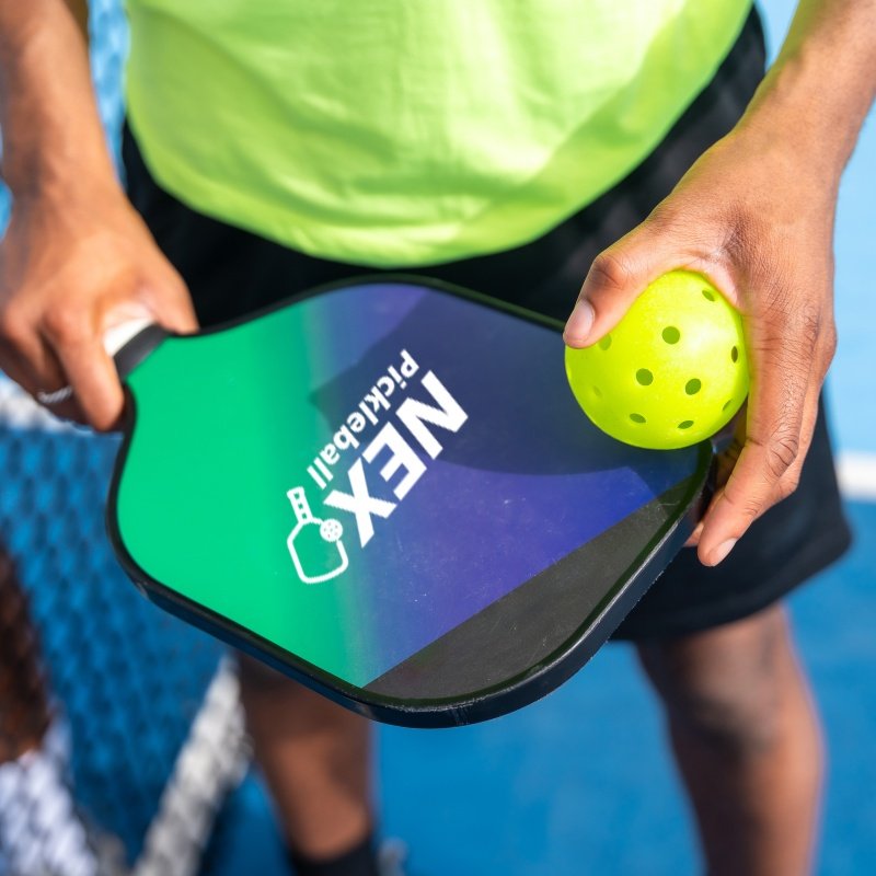 Close-up of hands holding a NEX Pickleball paddle and a yellow pickleball on a court.