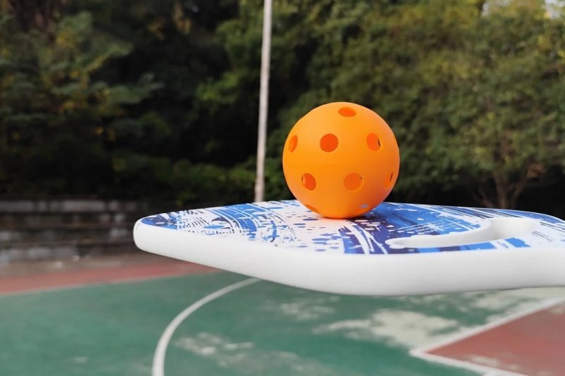 Pickleball and paddle on a vibrant outdoor court Orange pickleball resting on a blue and white paddle on an outdoor court
