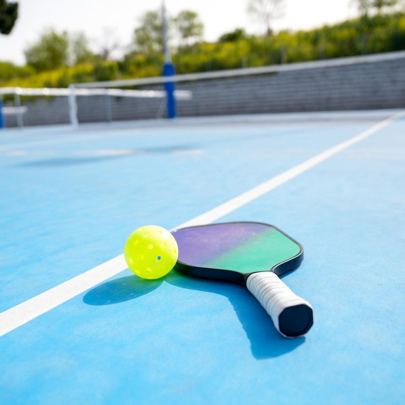 Pickleball paddle and yellow ball placed on a blue court under sunlight.