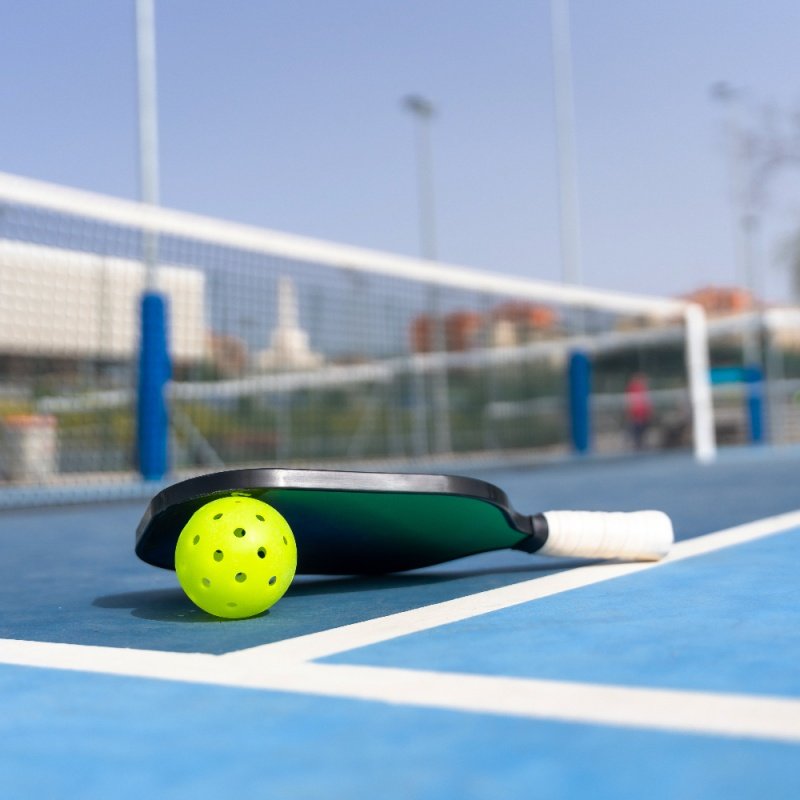 Pickleball paddle resting on a blue court with a yellow ball near the net.