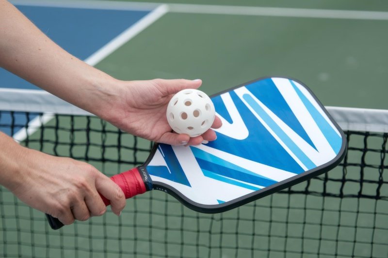 Player holding a white pickleball and blue paddle near the net on a court
