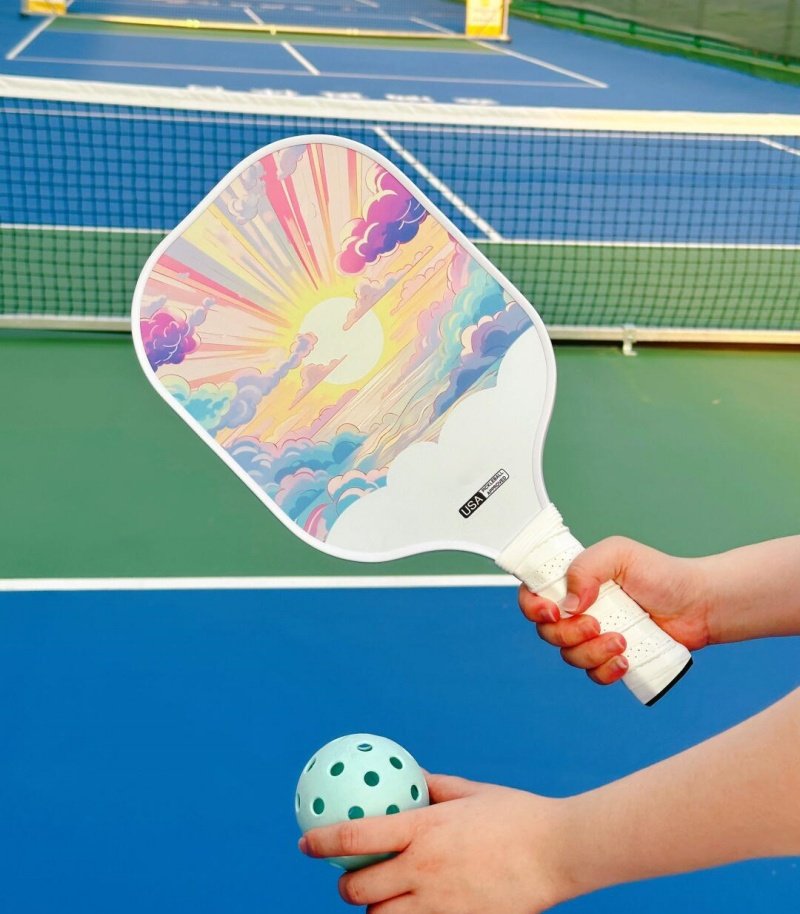 Pickleball paddle with vibrant cloud design and teal ball Pickleball paddle with colorful cloud design held by a hand, with a teal ball in the other hand