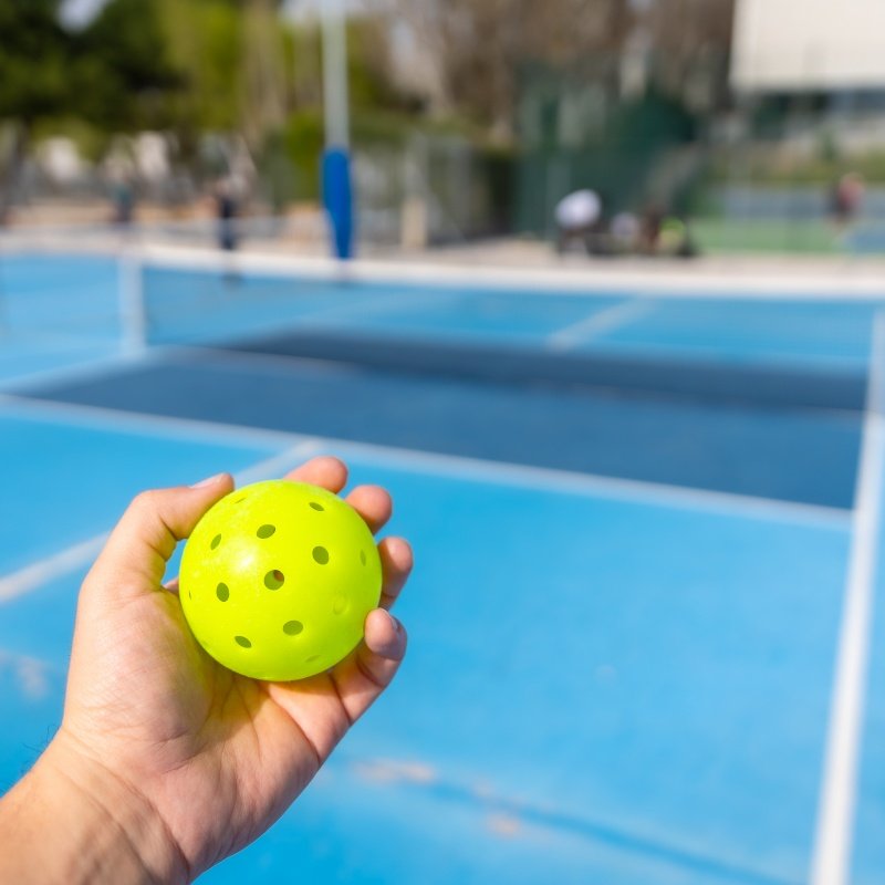 Hand holding a yellow pickleball ball with a blue outdoor court in the background.