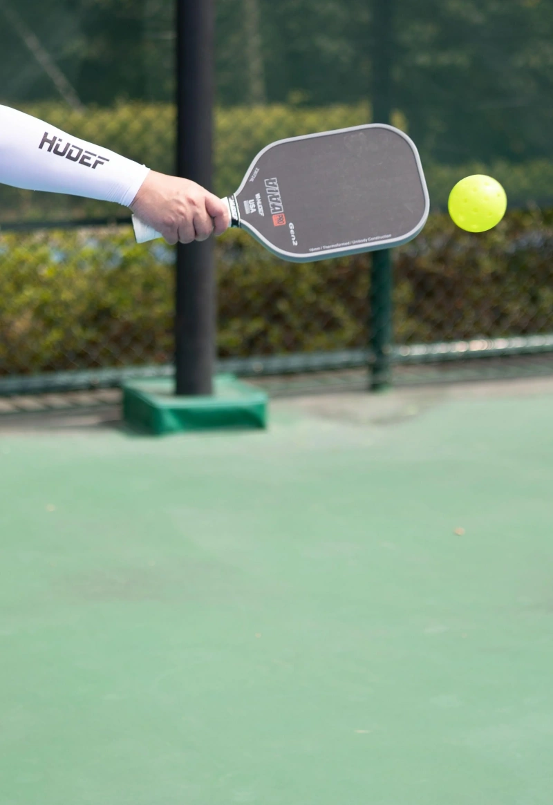 Hand holding pickleball paddle with a yellow ball in the air on court