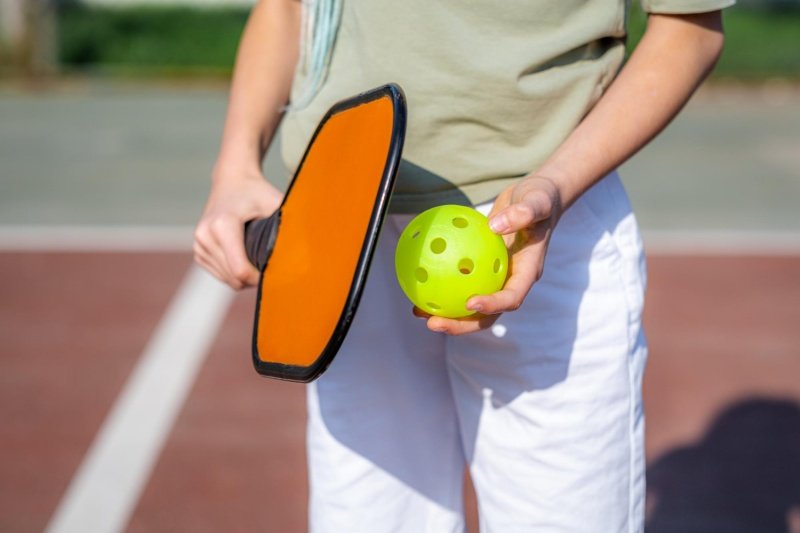 Person holding orange pickleball paddle and yellow ball on court