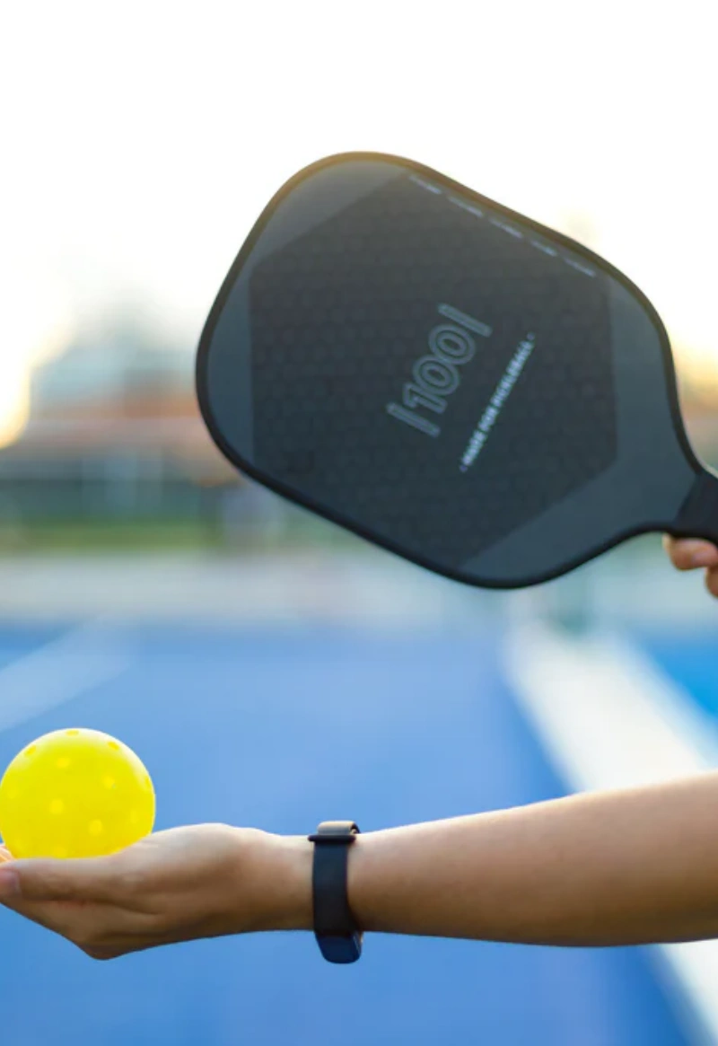 Hand holding yellow pickleball ball and paddle in the background of a court