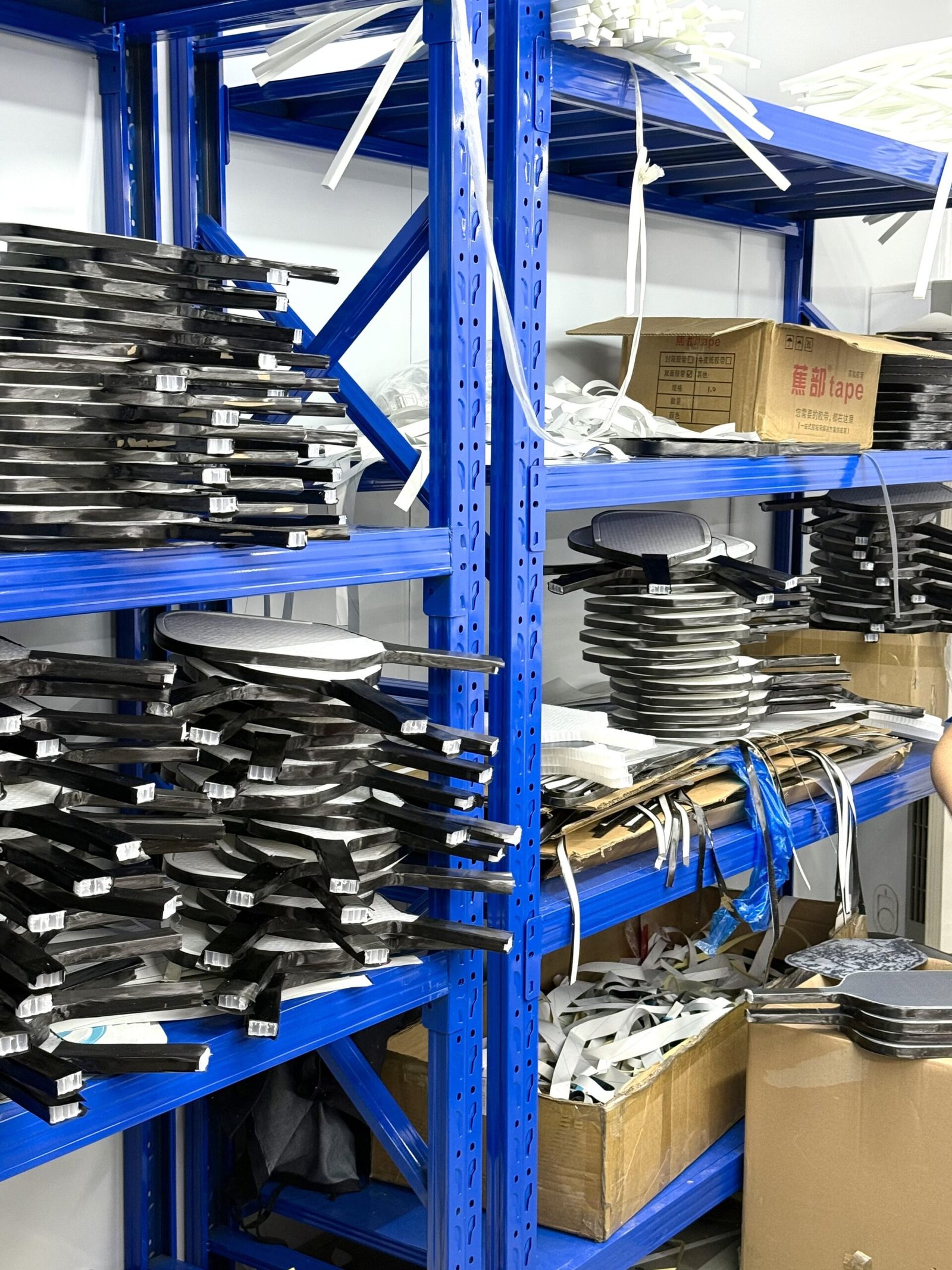 Paddle Storage Area Unfinished pickleball paddles stacked on blue metal storage racks