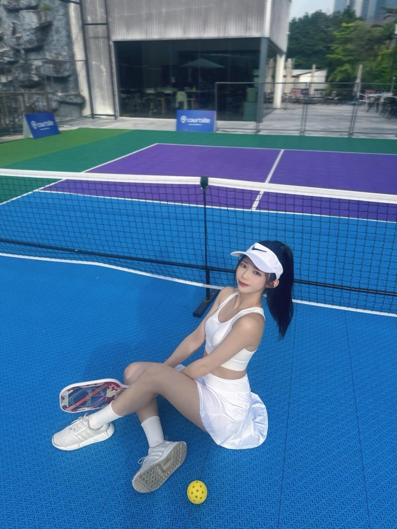 Pickleball Court Break Smiling woman in white sportswear sitting on blue pickleball court with paddle and ball