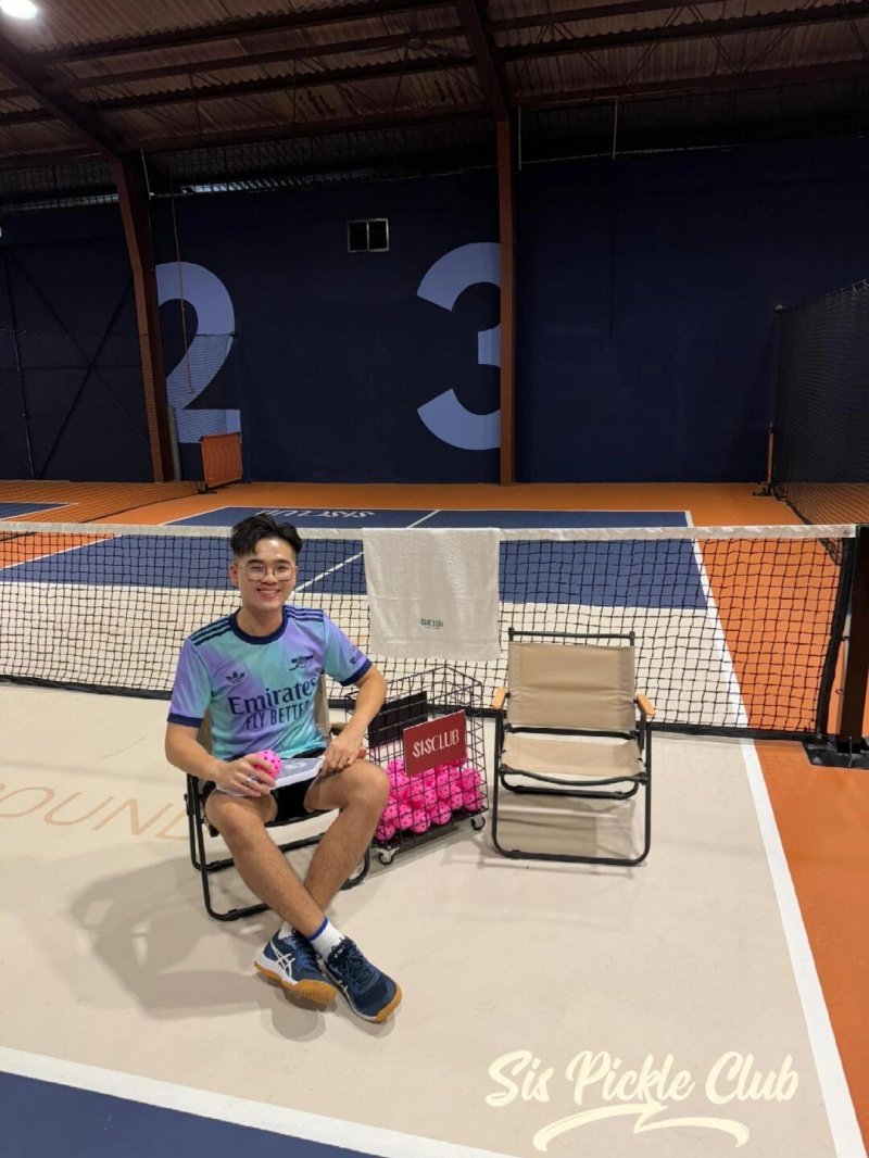 SIS Pickle Club Scene Young player seated beside basket of pickleballs at SIS Pickle Club court