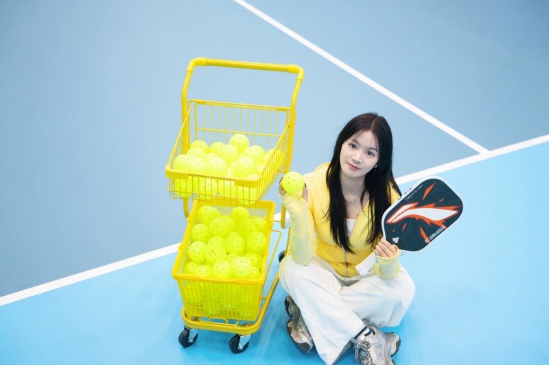 pickleball-cart-yellow-balls Woman holding a pickleball paddle and ball with a cart full of yellow balls
