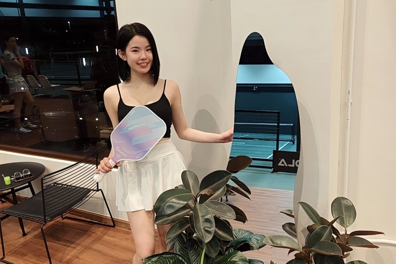 pickleball-indoor-smile Woman holding a pickleball paddle standing by plants with a court in the background