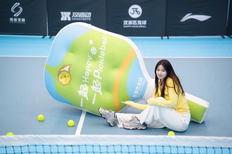 pickleball-inflatable-bottle Woman sitting next to an inflatable pickleball bottle with yellow balls on the court