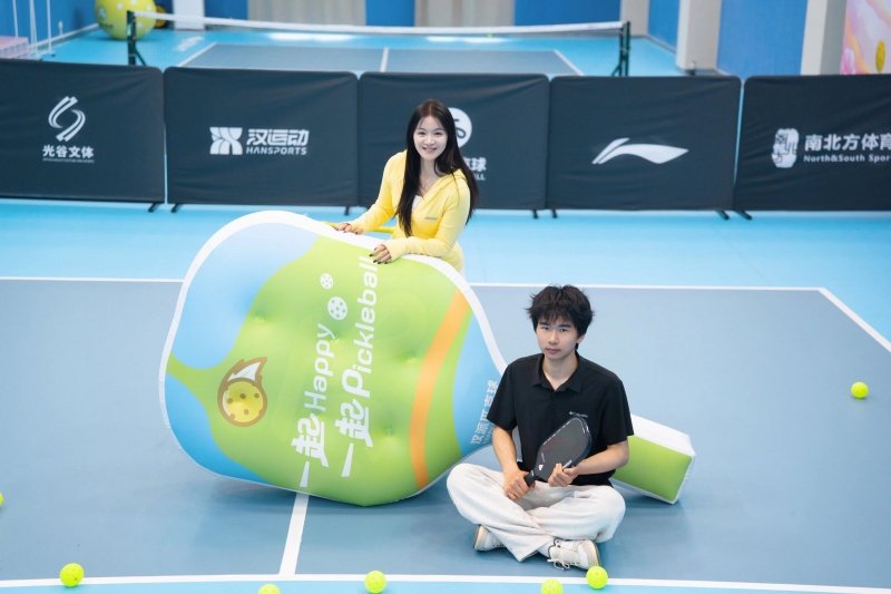 pickleball-inflatable-court Woman and man sitting next to an inflatable pickleball bottle with balls on the court